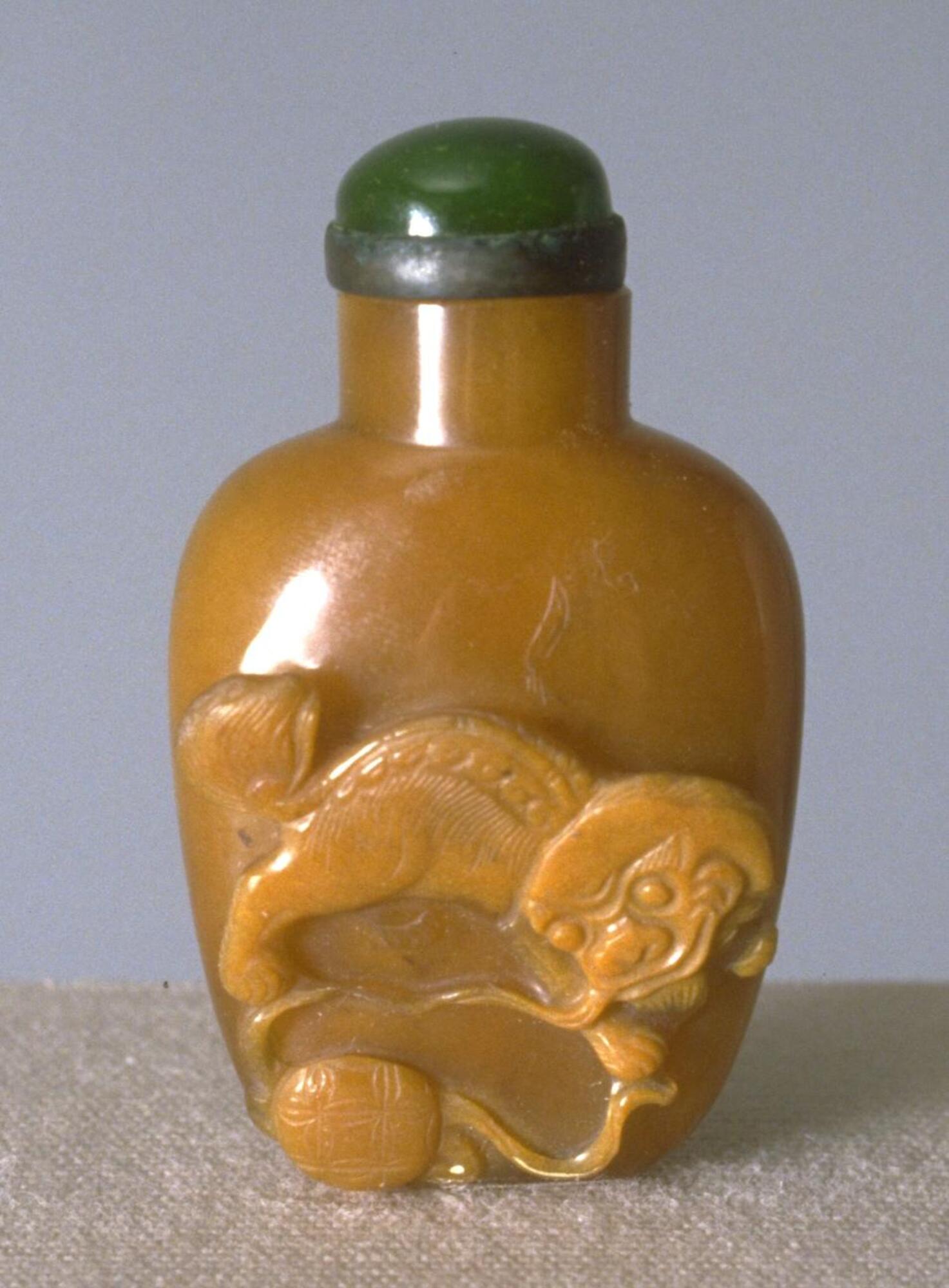 An orange agate snuff bottle with the image of a lion and a ball carved in raised in raised relief. On top of the snuff bottle is a green glass stopper in a collar.
