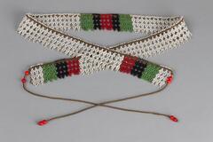 Beaded necklace in lattice pattern. White beads on brown thread with three sections of green, black, and red beaded stripes. Long threads at end with four red beads on each. Threads are 18 inches long.