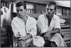 Two prisoners in uniform sitting on a wooden bench. Their legs are crossed and they are both looking to the right.