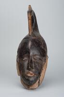 Wood mask of an elongated face topped with a bird head and beak.