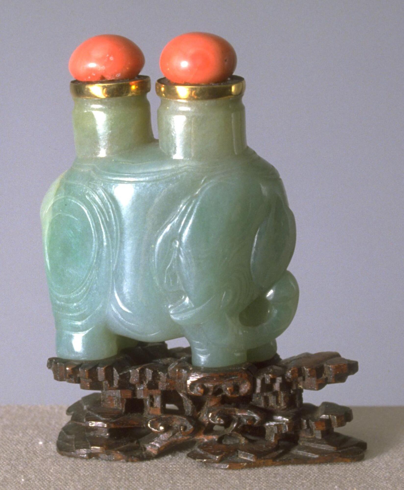 A bluish-green nephrite jade snuff bottle in the shape of an elephant with its head positioned to the side. On the back of the elephant are two openings with gold rims and oval coral stoppers. &nbsp;Carved on the sides of the elephant are &nbsp;circles that show the creases on the elephants body.