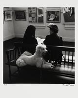 Two women sit on a bench in an art gallery, backs to the camera. A light-colored poodle sits on the bench behind the women, leash falling to the ground.