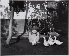 Photograph of a woman sitting outside a house with a bowl in her lap and ducks at her feet. 