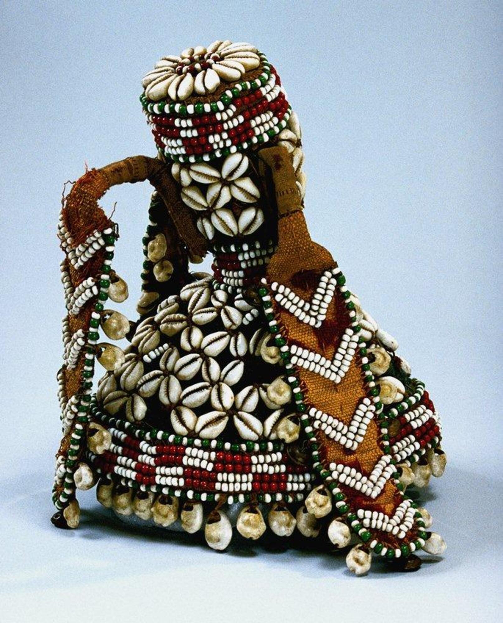 A cone-shaped headdress with a cylindrical finial rising from its top. Three long flaps hang from three projections located near the top of the finial. The green field of the main body and finial is embroidered with cowrie shells arranged in a four-pointed rosette pattern. Cowries also appear on the round flat top of the finial where they radiate around a circle of alternating red and white beads. Cowries form a fringe around the lower border of the cap and are accompanied by brass bells along the edges of the flaps. The headdress is bordered at its lower edge and at the top and bottom of the finial with a checkerboard pattern in red and white beads between thin rows of alternating green and white beads. Five double rows of white beads form a chevron pattern on the wheat-colored field of the flaps.
