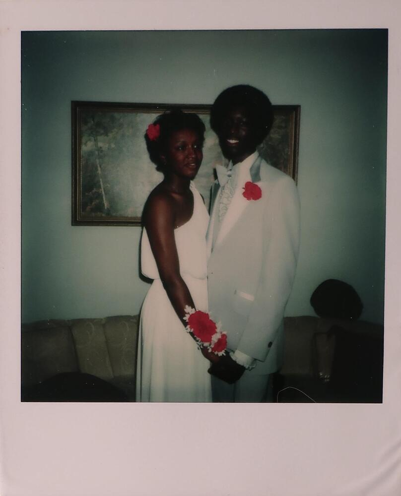 A young woman and man dressed in formal clothing, posing together in what appears to be a living room. The woman wears a white one-shoulder dress with a red flower in her hair and a red flower corsage. The man wears a powder blue tuxedo with a red flower on his lapel.