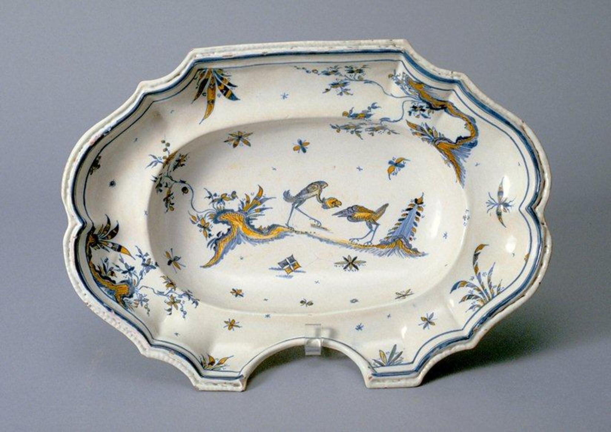 This oval-shaped ceramic vessel features a wide, steep rim surrounding a large well in the center. The scalloped edges of the rim are cut back into a deep semicircle on one side. The rim is painted in underglaze blue and yellow with plant and floral motifs that are arranged in a whimsical, asymmetrical fashion around the edge. The well is decorated with similar vegetal motifs and two vaguely stork-like birds with long legs that confront one another on a strip of turf.