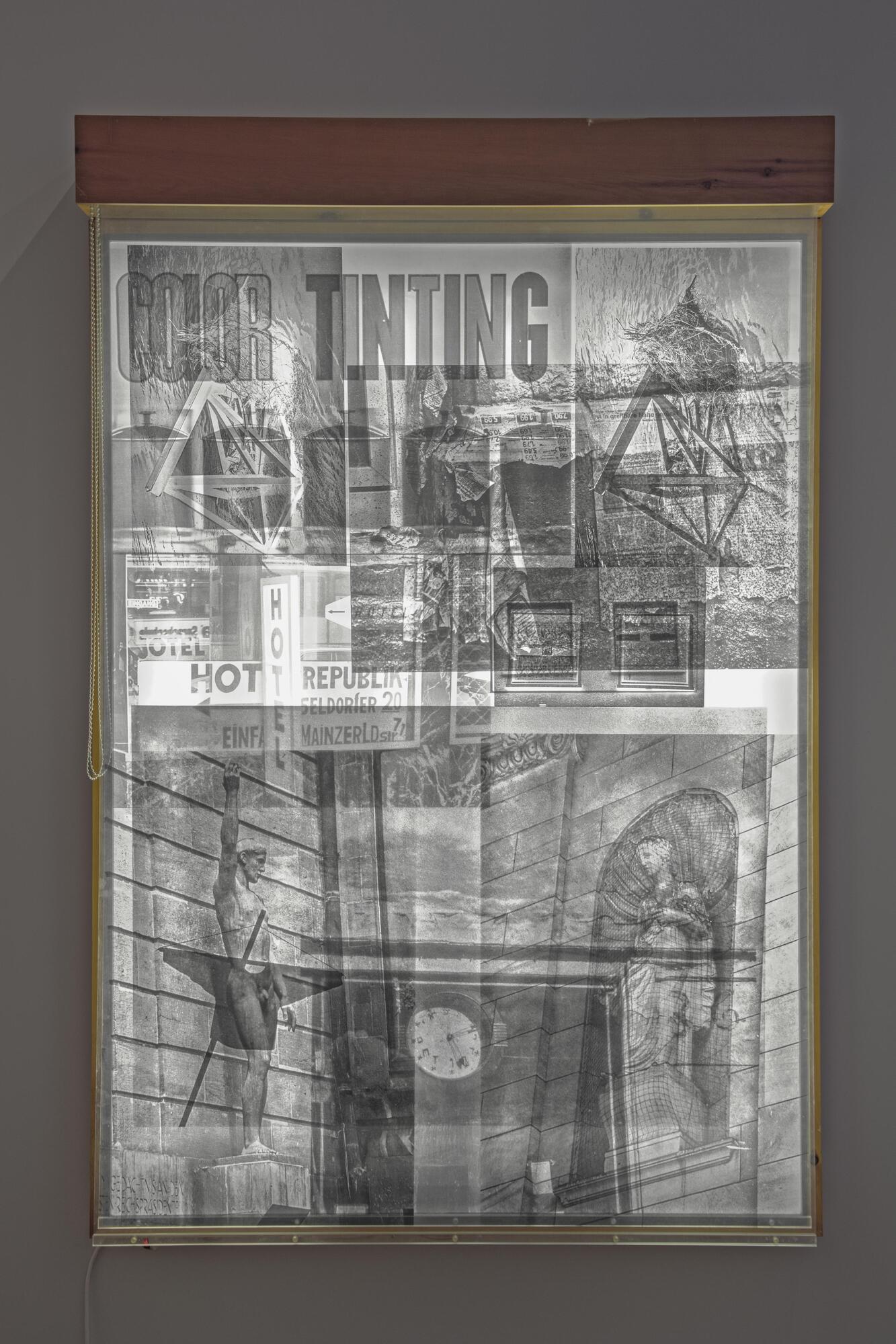 This mixed media assemblage has a series of see-through panels hanging from a wooden frame with a lightbox at the back. On the hanging panels, there are photographs printed, with a variety of images of buildings, sculpture, and advertising.