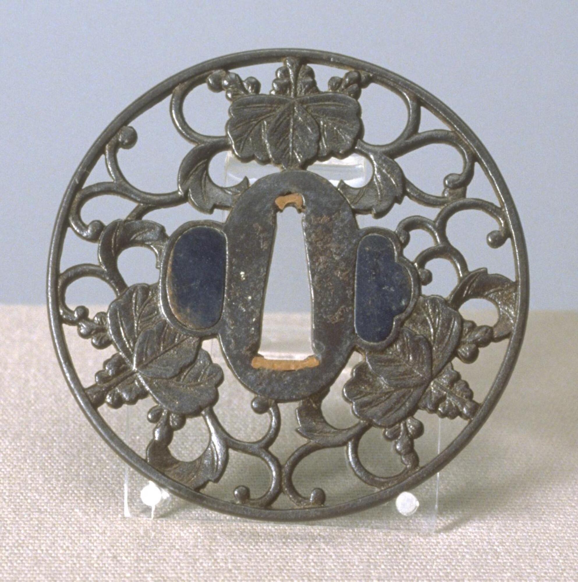 This small, flat metal piece has a circular shape and an openwork design. It has a triangular shaped sword hole in the center, flanked by two other holes, which are filled with shakudô (copper-gold alloy). The sword hole is mended with gold. Three crests, consisting of pawlownia leaves and flowers, are interconnected with vines. There are some abrasions on the center oval shape around the sword hole. The surface is slightly textured by minute stippling. The outer rim is slightly elevated from the inner design. This openwork carving technique is called "marubori" (round carving).