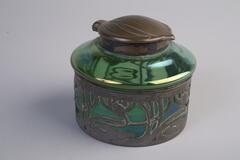 A circular green glass inkwell with a metal overlay on the bottom half. The overlay is a floral pattern and the lid is shaped like a leaf or flower petal.