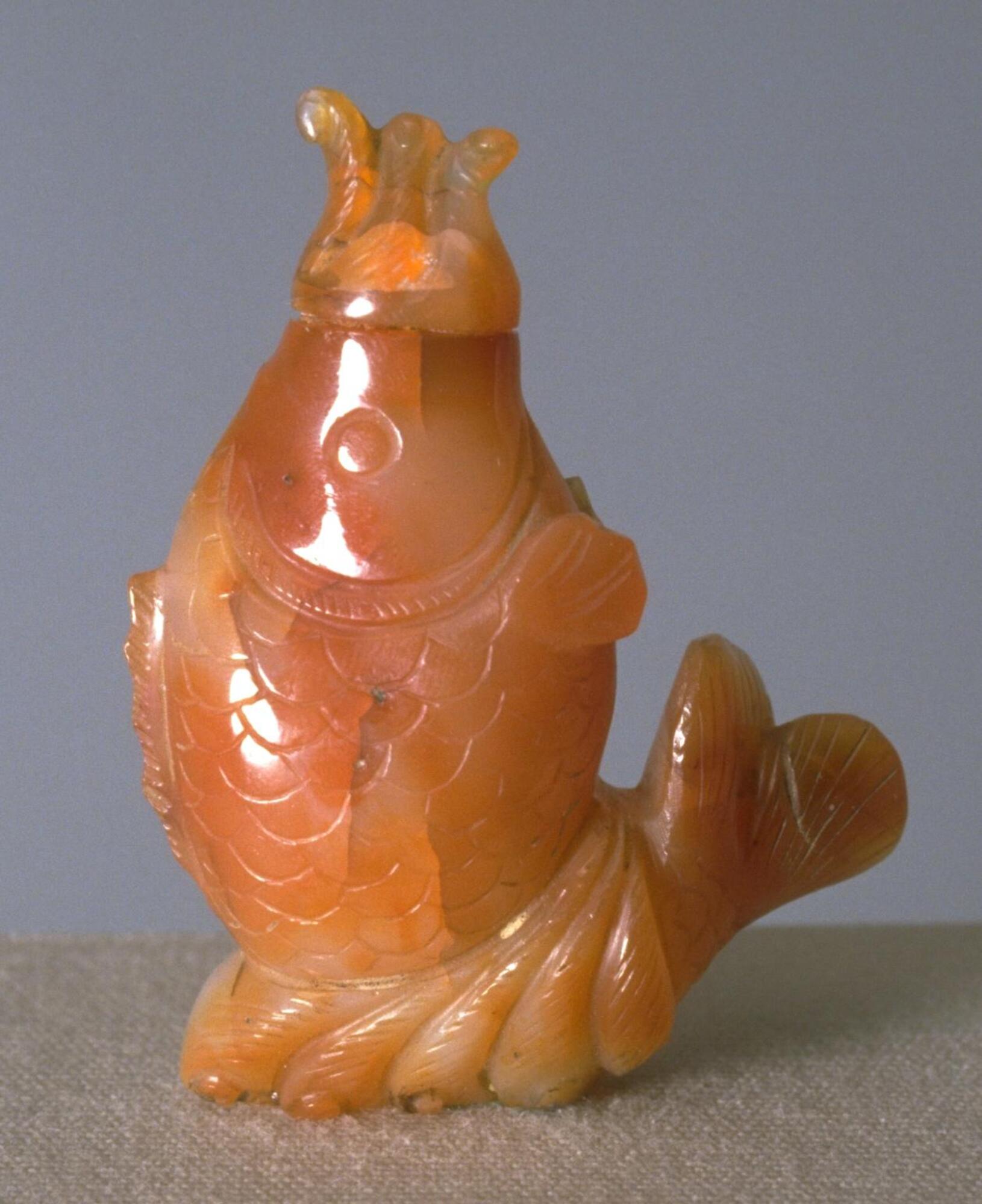 A carnelian snuff bottle in the shapes of a carp. Its head is raised and in its mouth is a stopper.