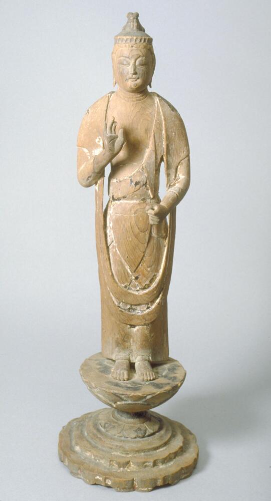 The figure is standing on a lotus-shaped pedestal; the hair is tied as a knot on top of the head; a crown is also on the top. The face has two elongated ears, round eyeblows, eyes looking downward; the lips are shut; sloping sholders are wrapped with thin robe, which hung toward the knees. Right hand, showing a palm, is raised to the chest while the left hand is by the lower abdomen, as if holding something. The three wrinkles can be seen on the neck. All are made of wood.