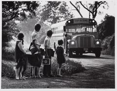 Kids waiting for a bus. One boy with his hand on a dog while they stand there. The bus is coming down a dirt road.