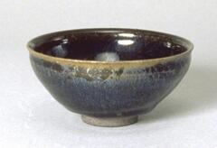 A deep, conical bowl on a straight foot ring with subtle rim articulation, covered in a thickly applied, dark, iron-rich black glaze with silvery-blue hare's fur markings (兔毫盏 <em>tuhao zhan</em>). The thick glaze thins at the rim to a lighter caramel-brown color.