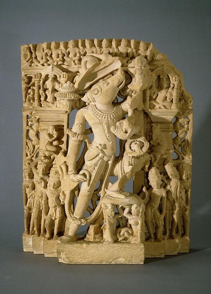 This intricate stele has a large Varaha in the center.  He is in the archer’s stance, with his right leg extended and his left leg bent resting on a lotus held up by a male and female snake figures. They have human bodies from the waist up and knotted snake bodies below.  Varaha has a human body with the head of a boar, his head thrown back supporting the figure of the earth goddess who holds on to his snout.  A lotus leaf acts as an umbrella over his head.  Three of his four arms are intact with his right one at his hip holding a broken lotus, only the stem survives, and the two left hands holding a conch at his chest and a discus at his knee.  The broken arm held the club and the top of it is still visible next to the pavilion on the left over his shoulder.  Besides the two snake figures, three figures stand on the base to either side, the other one female, while the others are male.  The inner two hold the conch and discus and can be considered shankhapurausha and cakrapurusha, the personifications of the 