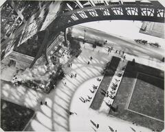 A black and white photograph of the area beneath the Eiffel Tower. A shadow from an arch of the structure covers most of the ground. A crowd of people stand beneath the tower.
