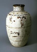 This tall, ovoid jar tapers to the base from slightly wider shoulders. Its wide neck has a slightly flared rim. The jar is covered in a white slip, then painted with dark brown-black calligraphic and free-form decoration, which is applied in rows between wavy bands around the body. There are a series of straight and wavy bands around shoulder and neck. The jar's mouth has a dark brown rim, and the entire form is covered in a clear glaze. 