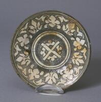 This ceramic plate contains negative white designs of a cross at center surrounded by a band of floral motifs at the rim. The plate is a gray-white porcelain whit glossy glaze and wide crackle. The colors used are primarily gray and white. The object was fired upright and is slightly restored. It probably dates to the Shah Abbas Safavid period. 