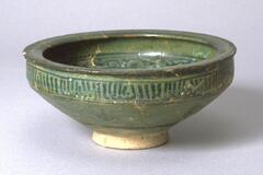 This dark green flat-rimmed bowl comes from the 13th century Iran and is decorated with abstract and painted designs. The interior features a floral design with buds, white the exterior has highly conventionalized inscribed bands and some floral elements.<br />
 