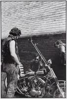 A photograph of three men gathered around a motorcycle. A man in the foreground stands with his back to the camera, while the two other men examine the machine. Behind them, a brick wall is partially illuminated by bright sun.