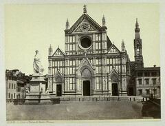 This photograph depicts a view of a church. Before the church in the piazza stands a monumental statue of a figure in draped clothing.  