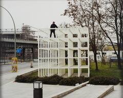Color photograph of a man standing in a cube-shaped structure made of white slats in an urban area.
