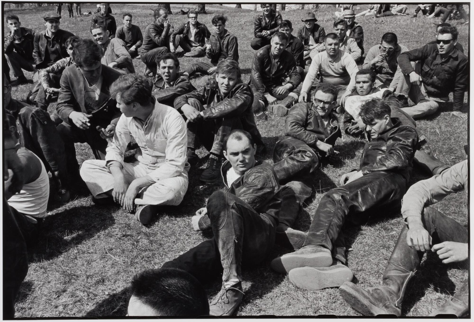 A photograph of a large group of men on a grassy lawn. Many wear leather jackets, jeans, and recline or sit cross-legged.