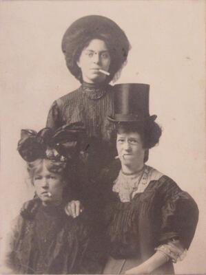 A portrait of three women in dark-colored, Victorian-style dress. The two in front are seated and the third stands behind them with a hand on each of their shoulders. All three have cigarettes in their mouths and are looking directly at the camera.