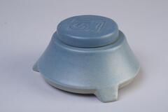 An inkwell made with ceramics in blue color. It is flat and round, wider on the bottom, and narrower on the top; and the lid cover has a ram head crafting on it.