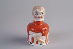 Inkwell made of porcelain has a cylinder shape body. The body of the inkwell is shaped to be a body of a woman cook, in orange clothings and a pinroller on her hand; the woman's head is the lid cover.