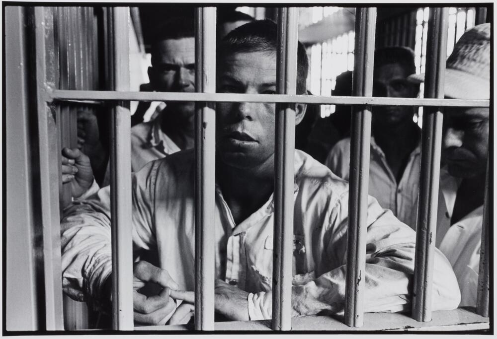 A man with his arms crossed, resting on the horizontal bar of a cell. There are other prisoners behind him.