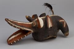 A wooden mask with zoomorphic features. The mouth is in the form of elongated lips, resembling an open beak. Each eye is formed by a short rod in front of a small rectangular projection. Behind the eyes are triangular ears. At the back of the head are two curved projections. The mask is painted red, white and black with a brown feather at the top of the head. 