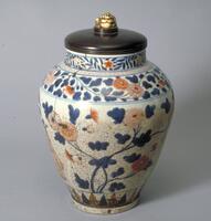 A medium size, well potted porcelain jar with wooden rid, round shoulder and neck. Floral designs are painted with blue underglaze and red and gold overglaze enamels. There are Chinese scholar and attendant boy with a fan on one side and Japanese lady in kimono on the opposite side, painted with enamels. Band of flowers on the neck, another broader band of chrysanthemums on the shoulder. There is also a band of leaf patterns on the bottom. A large crack from neck to the middle of the body; porcelain glaze has small cracks all over the body. The foot is unglazed; the eye is fully glazed. No glaze on the rim. The teak wood lid, a later addition, has a finial made of an ivory netsuke of laughing Hotei.