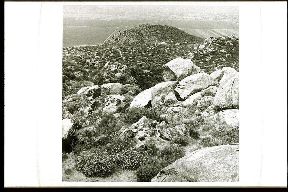This is a black and white photograph of an outdoor scene of a rocky hilltop. In the foreground are large boulders with vegetation growing among them. The hillside slopes down toward a distant valley iwith cultivated fields and straight roads. On the horizon is another set of rocky hillsides.