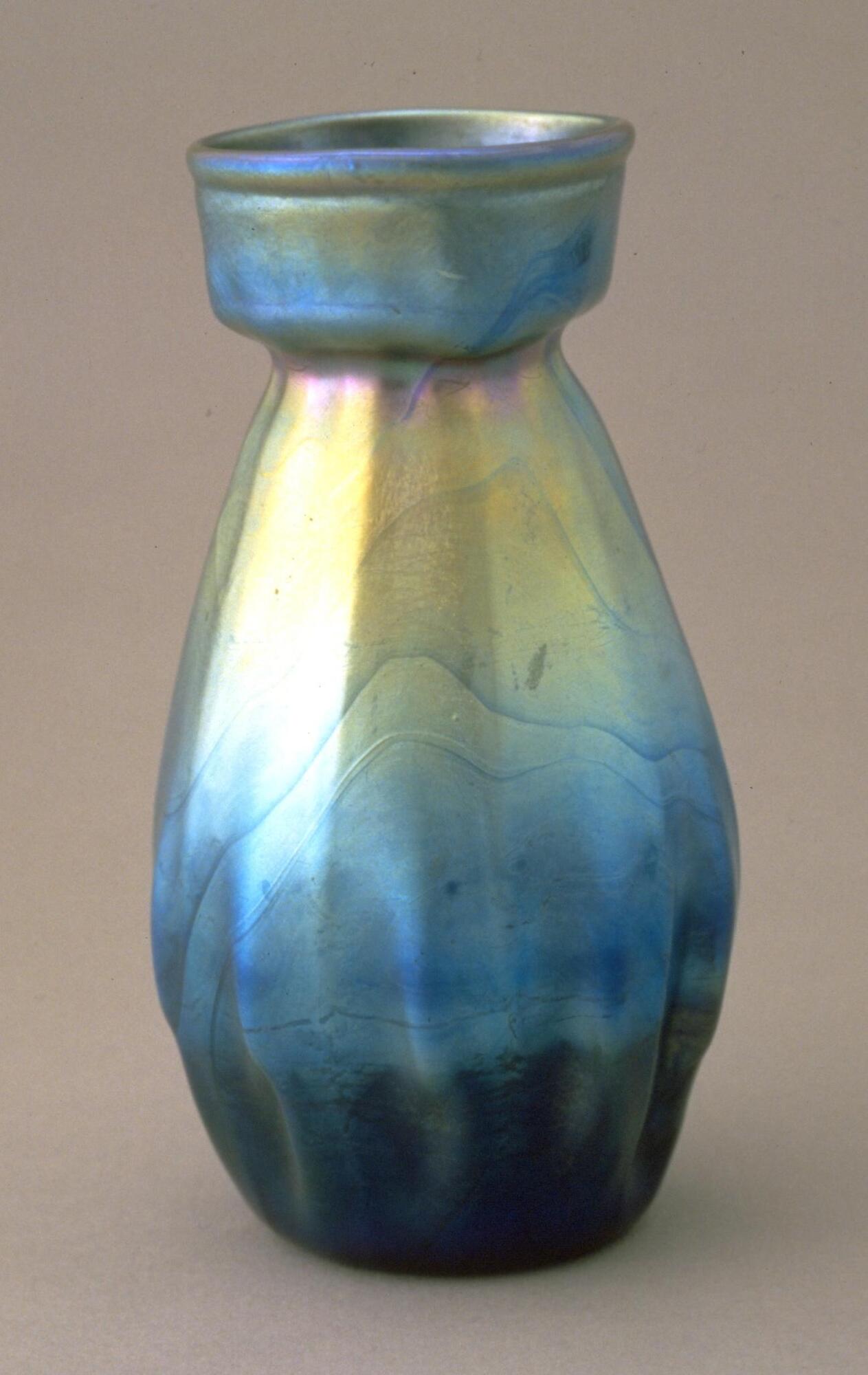 Iridescent blue, silver and yellowish-green glass vessel with egg-shaped body and the suggestion of lobes near the bottom half of the vessel tapering into eight flat sides towards the upper half. Vessel is topped by a soft-edged open-ended cube-like form.