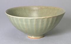 A conical stoneware bowl on a tall foot ring, the exterior wall fluted, covered in a craqueleur gray-green celadon glaze. 