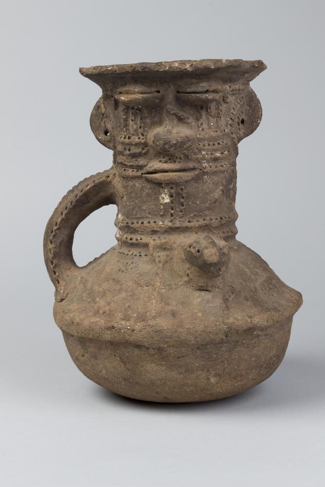 Clay vessel with a rounded bottom and a tall neck and handle. The bottom part of the vessel and handle are decorated with small holes and geometric patterns. The neck of the vessel is decorated with a face on both sides. The lips appear to be pursed and the eyes appear to be closed. The  half-circle ears stick out from the top of the neck, just below the rim of the vessel. On each cheek there are three horizontal marks with small holes. 