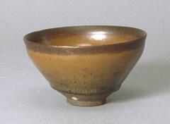 This deep, conical bowl rests on a straight foot ring with subtle rim articulation. It is covered in a thickly applied dark iron-rich brown-black glaze with hare's fur or <em>tuhao zhan (兔毫盏 ) </em>markings. The thick glaze pools in one black drip lowering onto the exposed base. The interior surface has a crackle glaze.