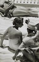 A sitting woman in a swimsuit turns her head to look at the man in a straw hat next to her. He wears a light shirt with a pattern of palm trees, raising his right arm in an unclear gesture.