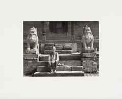 An old man sits off center on stone steps laid between two lion statues. The man wears a brimless hat, shirt, vest, and no shoes. The lions have a thick chain with a bell carved around each of their necks.
