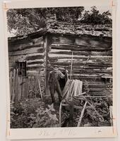 An old man wearing a hat, loose shirt, and trousers held on by suspenders stands outside a log cabin. He leans on a hoe with wood boards and agricultural tools nearby.&nbsp;
