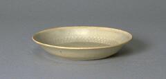 A thin, small porcelain plate with a flat bottom, flaring sides and a direct rim on a footring. The interior has an incised decoration of bands of lotus petals and a key fret pattern. It is covered in a white glaze with bluish tinge, with the rim unglazed.