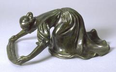 This bronze sculpture depicts a female figure kneeling, bending forward with her arms outstretched. She grasps a long piece of cloth that is stretched on the ground in front of her, as she looks directly at the cloth with intent. The woman is wearing a long skirt, which covers her legs and feet and swirls around her on the ground. Her blouse has flowing sleeves that end at the elbows, leaving her forearms bare. Her hair is pulled back and gathered into a knot. The bronze has a dark green patina and the surface of the statue is smooth and polished.