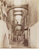 This photograph depicts a street-level view down a medieval Italian alleyway. The alleyway has archways that connect the buildings on either side. Seated on the street and leaning out of a window, a small group of spectators face the camera. 