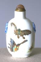 A white porcelain snuff bottle with painted designs of butterflies and relief of blue taotie masks. On the top is a glass stopper in a brass collar.