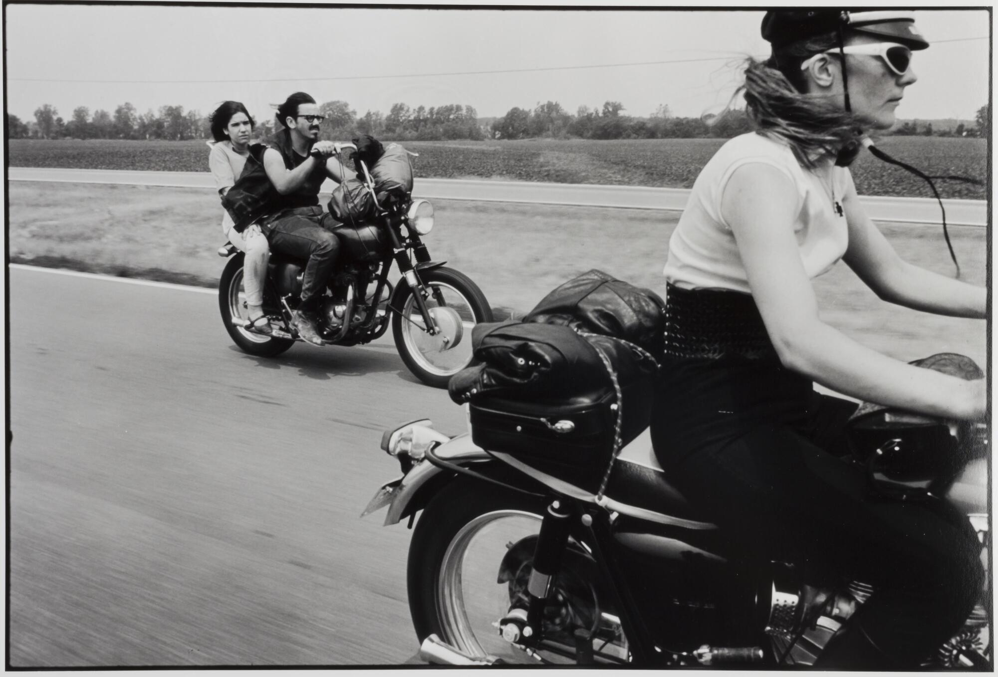 A photograph of two motorcycles driving on a highway. A woman rides the first motorcycle, which is cropped at the right-side of the frame, while a man and woman ride behind in the mid-ground.
