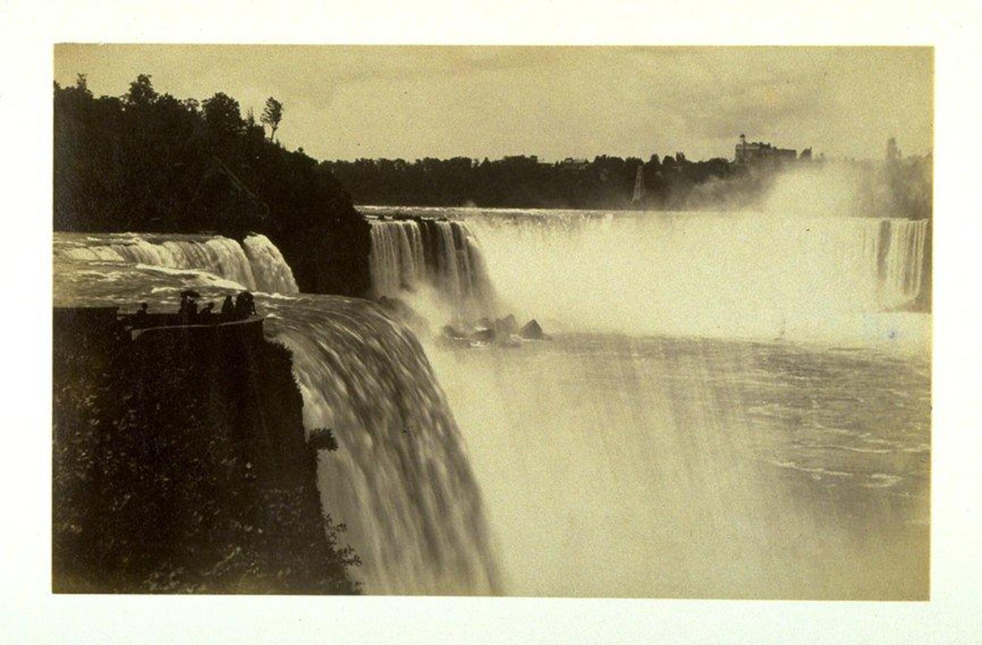 This photograph depicts a scenic view of Niagara Falls from a distance.  To the left of the image, a small group of people stand on an overlook.