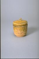 A thickly potted, cylindrical stoneware jar on a narrow footring.  The body is incised with a floral meander and topped with a flat cap lid with a loop finial.  The lid and half the body are covered in an amber-green glaze. 