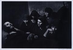 A group of women all wearing black with their heads covered in black mourning a man in a suit laying down. His hands are crossed over his body and he has a beard.
