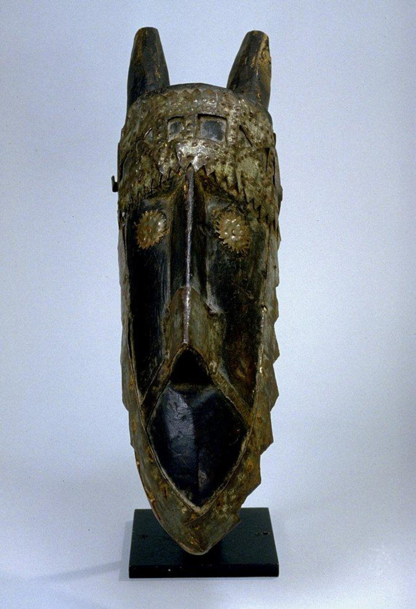 Stylized wood carving of hyena head;  two pieces of wood joined to form articulating jaw. edged with prominent teeth. short upright ears, long triangular nose or snout; eyes, teeth, nose and crown of head overlaid with sheet metal. Wood is unpainted.