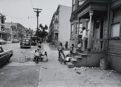 A dirty city street with row houses. A woman is on a porch drinking a coke. There are kids playing on the sidewalk with bicycles.